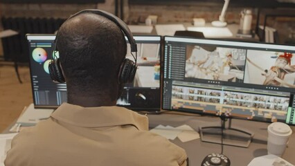 Medium closeup of male African American editor sitting at desk with computer, adjusting headphones and looking at screen in office in evening