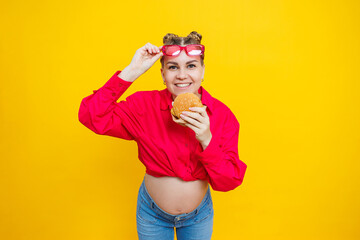 Close-up of a pregnant woman in a bright pink shirt and junk food. Hamburger and pregnancy. The concept of a pregnant woman eating unhealthy food. Cheerful pregnant woman eats fast food