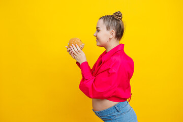 Funny pregnant young woman eating burger. Pregnant woman holding tasty sandwich while standing isolated on yellow studio wall. Junk food concept. Fast food during pregnancy.
