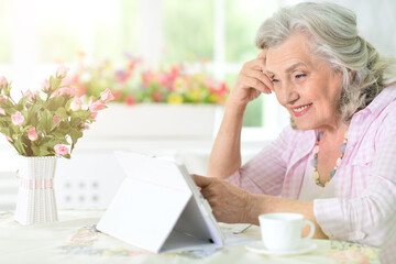 Beautiful old woman using modern tablet while drinking tea