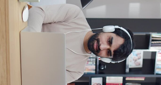 Vertical shoot. Focused young man wearing heaphones using laptop, typing on keyboard, writing email or message, chatting, shopping. Successful freelancer working online on computer.