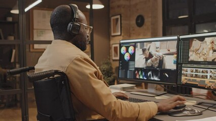 Tilt shot of male African American wheelchair user sitting at desk typing something on keyboard, using color grading control in office in evening