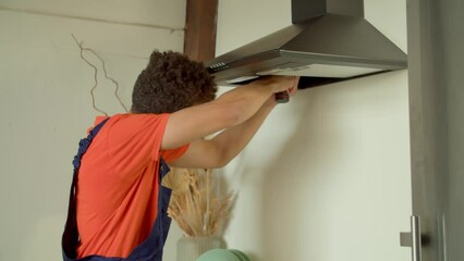 Portrait of professional attractive black male repairman in overalls making service appliance maintenance, fixing range hood with tools and installing household kitchen hood filters indoors.