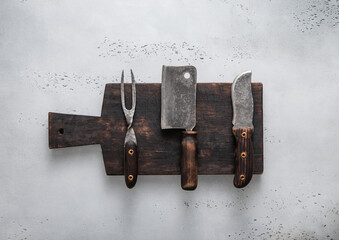 Chopping cutting board with meat hatchet and fork and knife on light background.