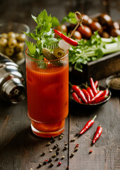 Bloody mary cocktail with red hot peppers and celery on wooden background with bar shaker.