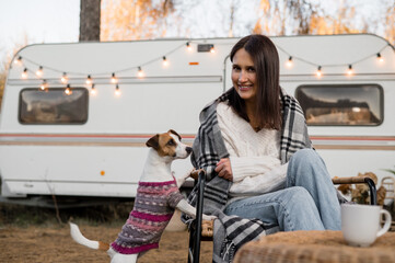 Caucasian woman sitting in a wicker chair wrapped in a blanket with a dog in the yard near the trailer in autumn. 