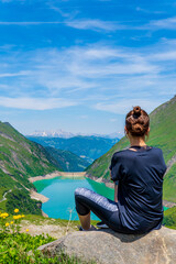 Fototapeta na wymiar Woman sitting and looking at Kaprun water reservoirs, Kaprun dam, Austria. Stunning turquoise picturesque lake surrounded by the green Alps mountains, blue sky, and clouds. Famous hiking destination.