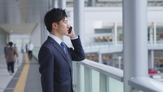 駅にてスマートフォンを使用して仕事をするスーツを着た若い日本人ビジネスマン