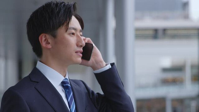 駅にてスマートフォンを使用して仕事をするスーツを着た若い日本人ビジネスマン