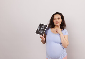 Emotional Latin American pensive pregnant woman, holding ultrasound scan of her baby, dreamily...