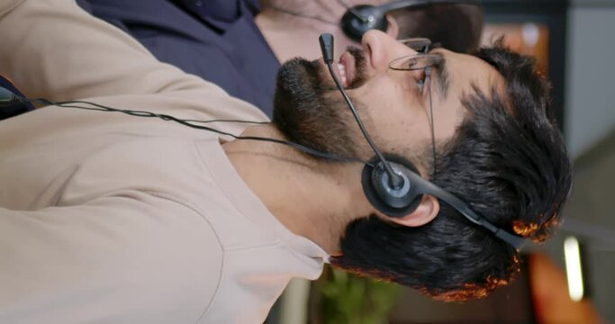 Vertical shoot. Adult handsome office worker wearing headhpones with microphone looking at camera and smiling. Berded man focused on work sitting at workplace. Operator of hot line.