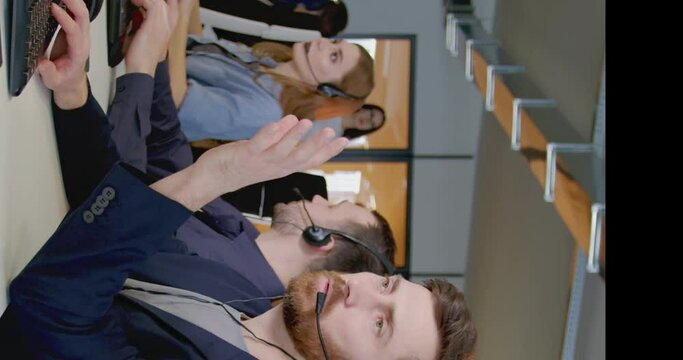 Vertical shoot. Adult european office worker wearing headhpones with microphone. Berded man sitting at workplace. Customer service representative with headset in call center. Consept of office work.