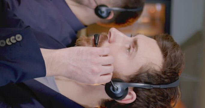 Vertical shoot. Adult european office worker wearing headhpones with microphone looking at camera and smiling. Berded man sitting at workplace. Operator of hot line. Consept of office work.
