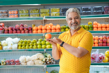 Senior indian man showing orange fruit at fruit shop. Healthy lifestyle concept.