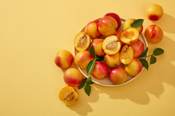 Colorful fresh ripe peaches (prunus persica) with green leaves on the stem are placed on a round plate and decorated on a yellow background. Minimal scene with copy space