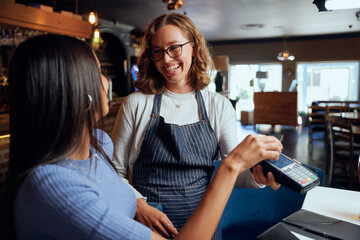 Happy young woman in casual clothing paying for dinner with card machine by waitress