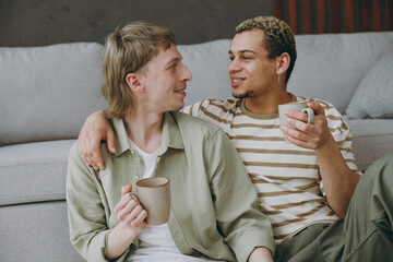 Young couple two gay men wear casual clothes together drink coffee look to each other sit on sofa couch at home flat rest spend free spare time in living room. Pride day june month love lgbtq concept.