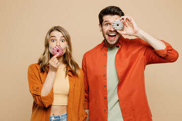 Young shocked surprised couple two friends family man woman wear casual clothes look camera cover mouth eye with donuts together isolated on pastel plain light beige color background studio portrait.