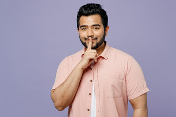 Young Indian man he wear pink shirt white t-shirt casual clothes say hush be quiet with finger on lips shhh gesture isolated on plain pastel light purple background studio portrait. Lifestyle concept.