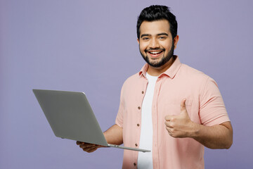 Young IT Indian man wear pink shirt white t-shirt casual clothes hold use work on laptop pc computer show thumb up isolated on plain pastel light purple background studio portrait. Lifestyle concept.