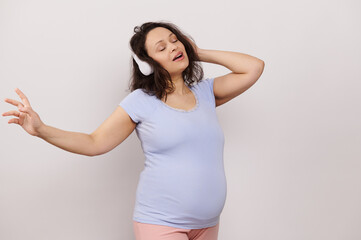 Multi ethnic cheerful delightful pregnant woman in blue t-shirt, singing and dancing while listening to soothing music on wireless headphones, posing with her eyes closed on white isolated background