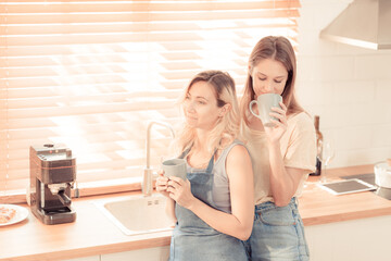 happy lesbian couple holding cups of coffee in kitchen. Couple of lesbian Girls Enjoy coffee at home taking about something. Two young adult beautiful women drink tea in modern kitchen. warm tone.