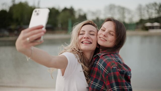Smiling hot girls enjoying themselves on the lakeshore during a pleasant summer evening. They are capturing moments with their mobile phones, taking selfies and photos to share on social media.
