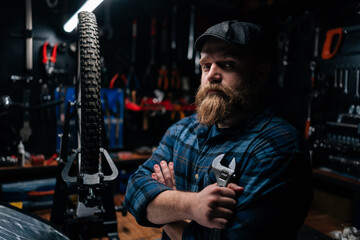 Fototapeta na wymiar Side view of bearded cycling repairman in cap holding wrench in hand standing by bicycle in repair workshop with dark interior, serious looking at camera. Concept of professional bicycle maintenance.