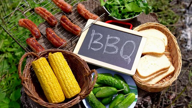 Upper view of tree stump with grill grate, grilled sausages, vegetables, corn, bread and cucumbers - summer backyard party. Girl hand lays down a chalkboard with the inscription BBQ. Video in 4k 25FPS