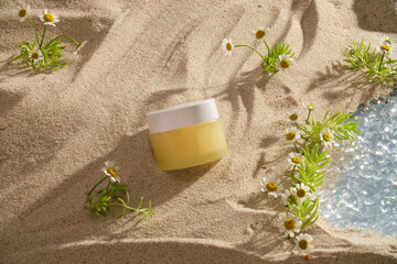Top view a cream jar displayed on sand background with flower, green grass and small lake. Mockup scene for advertising cosmetic with natural concept. Blank space for design