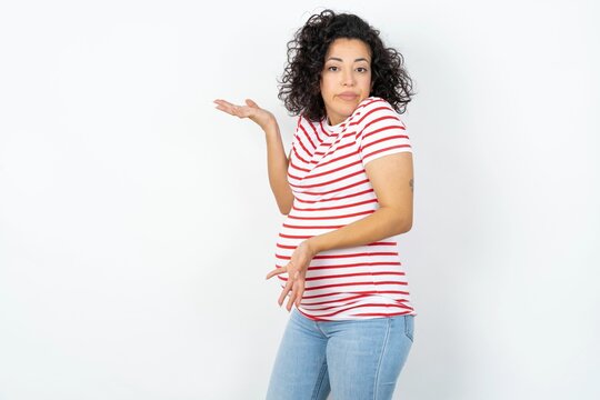 Beautiful pregnant woman wearing striped T-shirt standing over white studio wall pointing aside with both hands showing something strange and saying: I don't know what is this. Advertisement concept.