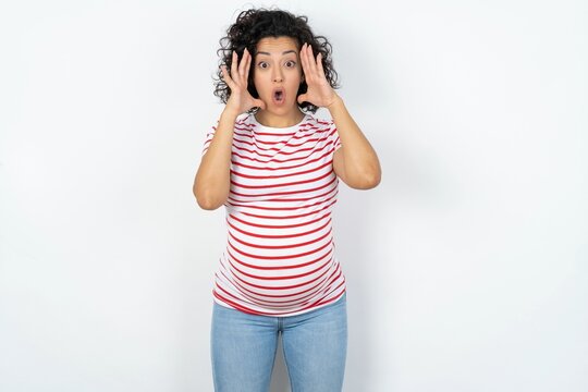 Beautiful pregnant woman wearing striped T-shirt standing over white studio background with scared expression, keeps hands on head, jaw dropped, has terrific expression. Omg concept
