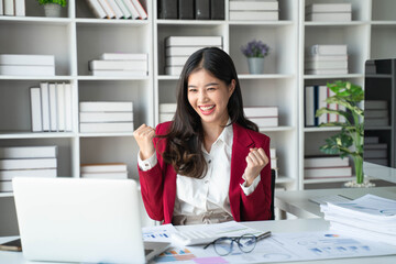 Asian business woman celebrating victory success Excited Success. Woman using laptop at work inside office holding hand up and happy triumph gesture.