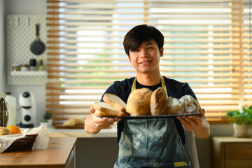 Happy handsome asian male baker in apron holding tray full of freshly baked and smiling to camera