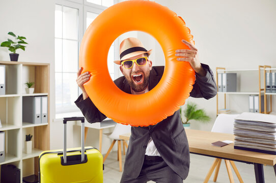 Happy funny crazy excited business man or office worker in suit jacket, sun hat and sunglasses standing in office, holding orange beach ring, looking at camera and screaming. Holiday, vacation concept
