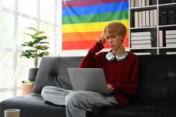Confused male student learning distantly, watching online lecture on laptop while sitting on couch with LGBT flag in the background