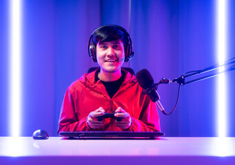 Professional gamer casting podcast walkthrough review. Young asian man sitting on chair with computer pc and microphone. Happy male Streamer wearing headphone playing game online in room neon light © Chanakon