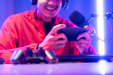 Professional gamer casting podcast walkthrough review. Young asian man sitting on chair with computer pc and microphone. Happy male Streamer wearing headphone playing game online in room neon light
