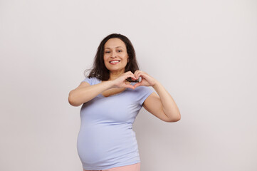 Obraz premium Beautiful pregnant woman smiling at camera, forming a heart shape with fingers, expressing happiness, positive emotions, enjoying her pregnancy and maternity lifestyle, isolated on white background