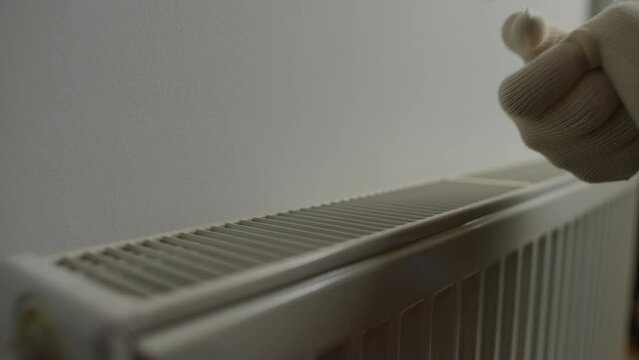 Closeup calefaction radiator and female hands in white gloves warming palms on the right leaving. Unrecognizable woman in cold apartment indoors