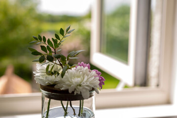 A mini bouquet of flowers in a jar on the windowsill of an open window to a green garden