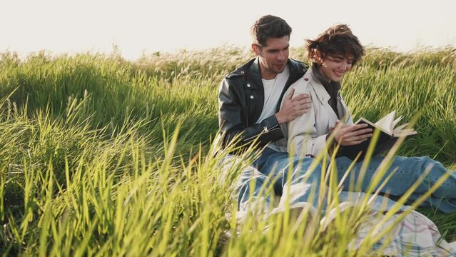 Smiling couple reading book and hugging while sitting on picnic blanket on field 