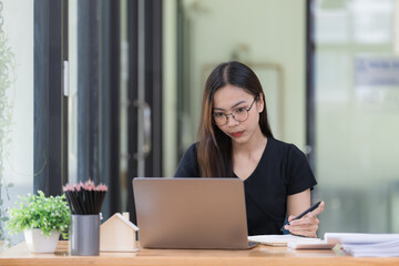 Fototapeta premium A beautiful young Asian businesswoman is saving and checking work and documents with her laptop computer at her desk with a bright smile on her face.