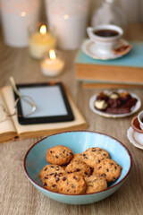 Books, reading glasses, e-reader, plate of chocolate pralines, bowl of cookies, cups of tea and lit candles on the table. Selective focus.