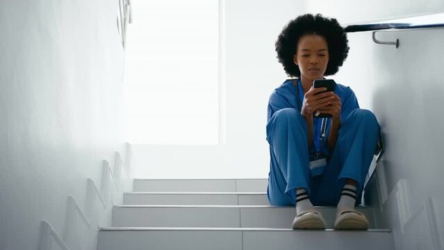 Female nurse wearing scrubs checking messages or scrolling online sitting on hospital stairs - shot in slow motion