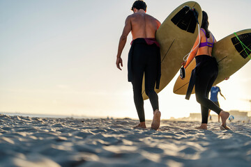 Rear view of couple of surfers carry surfboards on the beach. - Powered by Adobe