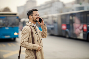 A happy urban businessman is standing on a bus station and having phone call.