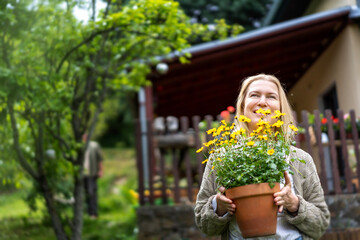 Happy middle aged blonde woman with a pot of flowers in her hands on the background of a country house. Summer lifestyle portrait