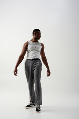 Full length of trendy young afroamerican man in sleeveless t-shirt and pants looking away on grey background, contemporary shoot featuring casual attire