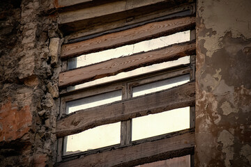 Fototapeta na wymiar Old window nailed with wooden planks of abandoned aged house with peeling plaster on bricks